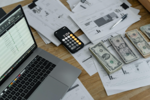 A laptop, a bunch of data files, money, and a phone on a work desk.