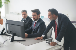 Three businessmen work on a dual-monitor computer.